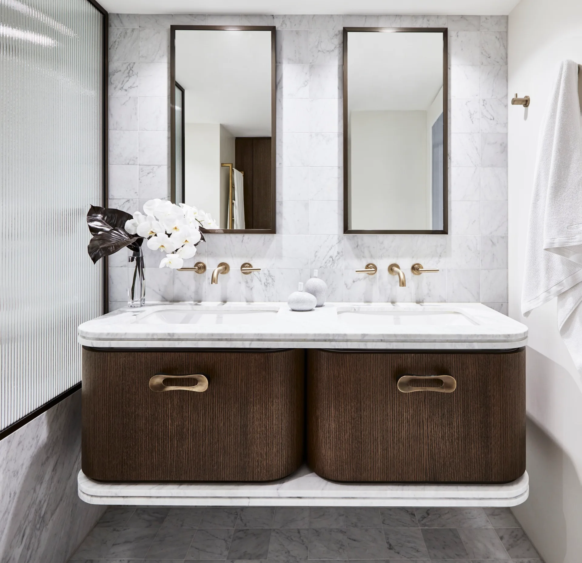 An ensuite bathroom with marble wall tiles, floor tiles and bath fascia. A glass screen blocks the view of the shower to the left. The timber vanity has a curved edge and marble benchtop. THe hardware is brass and two rectangle mirrors are positioned above twin basins set into the marble benchtops.