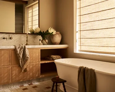 Modern farmhouse style bathroom with wooden beams, Roman blinds, a freestanding tub, and a vanity with flowers and a towel. The floor tiles are neutral with squares of brown and the vanity is timber. The result is a warm space with filtered sunlight.