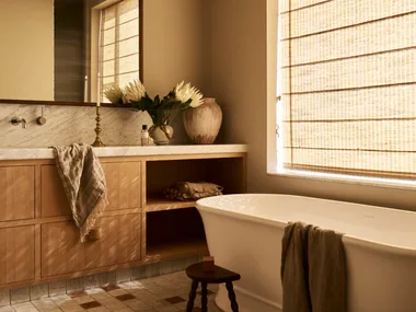 Modern farmhouse style bathroom with wooden beams, Roman blinds, a freestanding tub, and a vanity with flowers and a towel. The floor tiles are neutral with squares of brown and the vanity is timber. The result is a warm space with filtered sunlight.