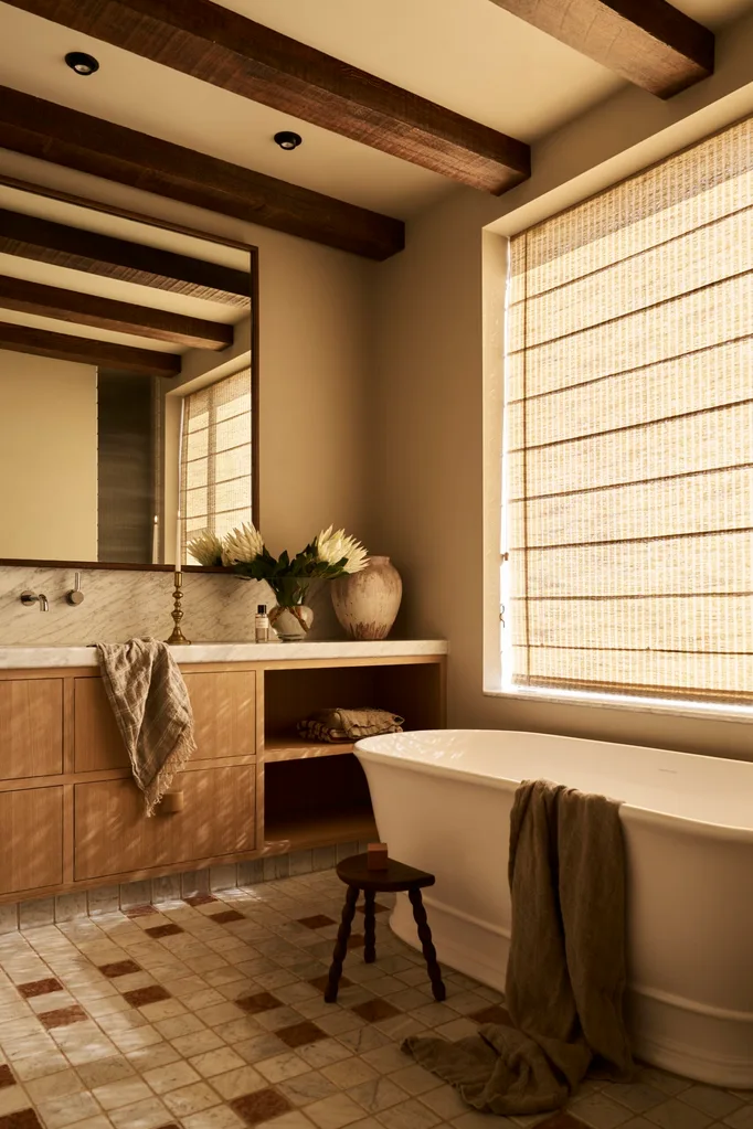 Modern farmhouse style bathroom with wooden beams, Roman blinds, a freestanding tub, and a vanity with flowers and a towel. The floor tiles are neutral with squares of brown and the vanity is timber. The result is a warm space with filtered sunlight.
