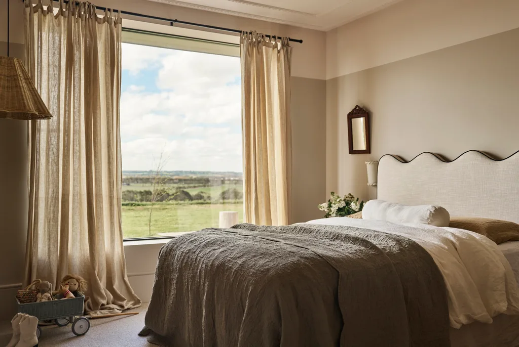 Cozy bedroom with a large window looking out to country views, white and gray bedding, beige curtains, wicker lampshade, and plush toys on the floor. The bedhead has a wavy shaped top.