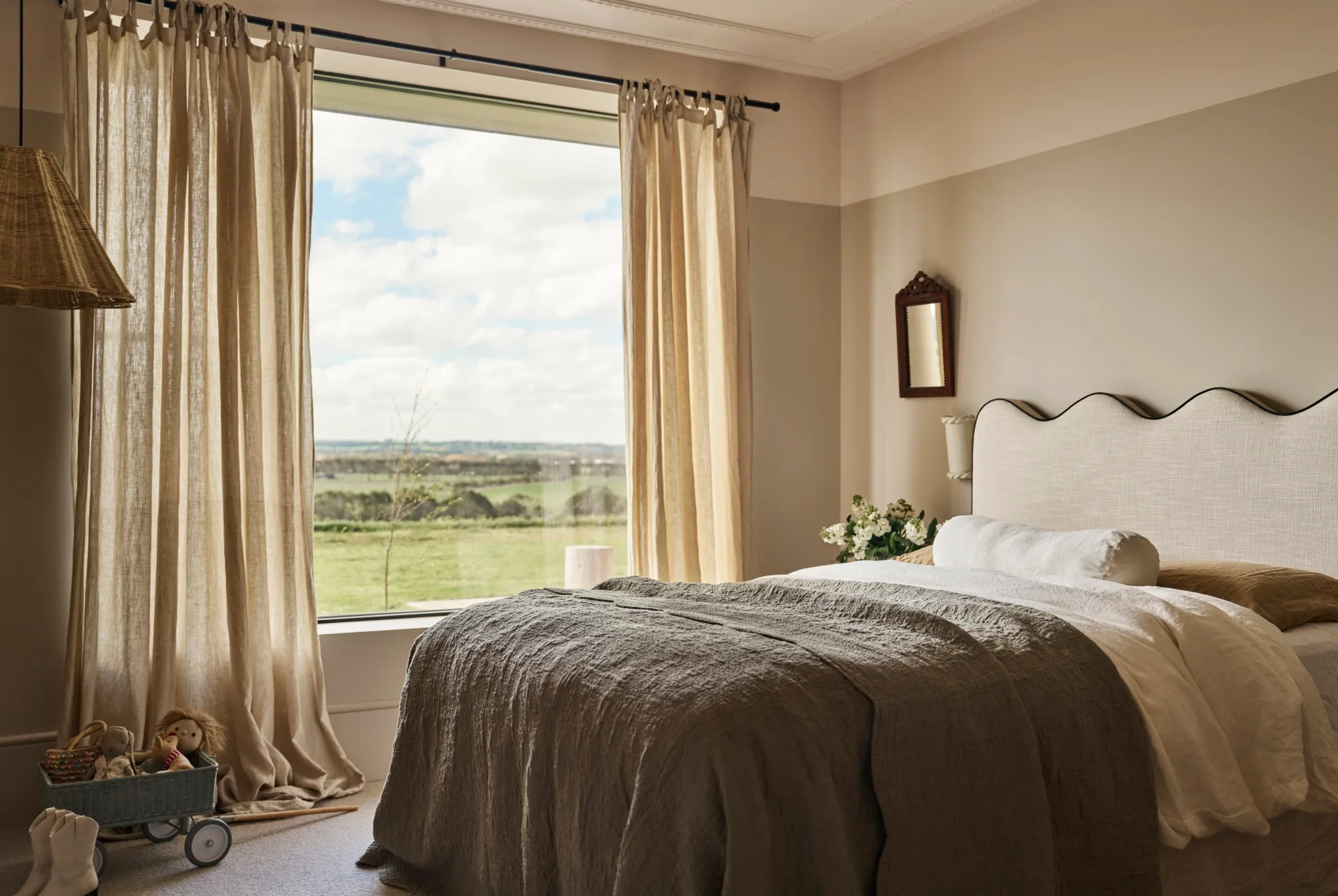 Cozy bedroom with a large window looking out to country views, white and gray bedding, beige curtains, wicker lampshade, and plush toys on the floor. The bedhead has a wavy shaped top.