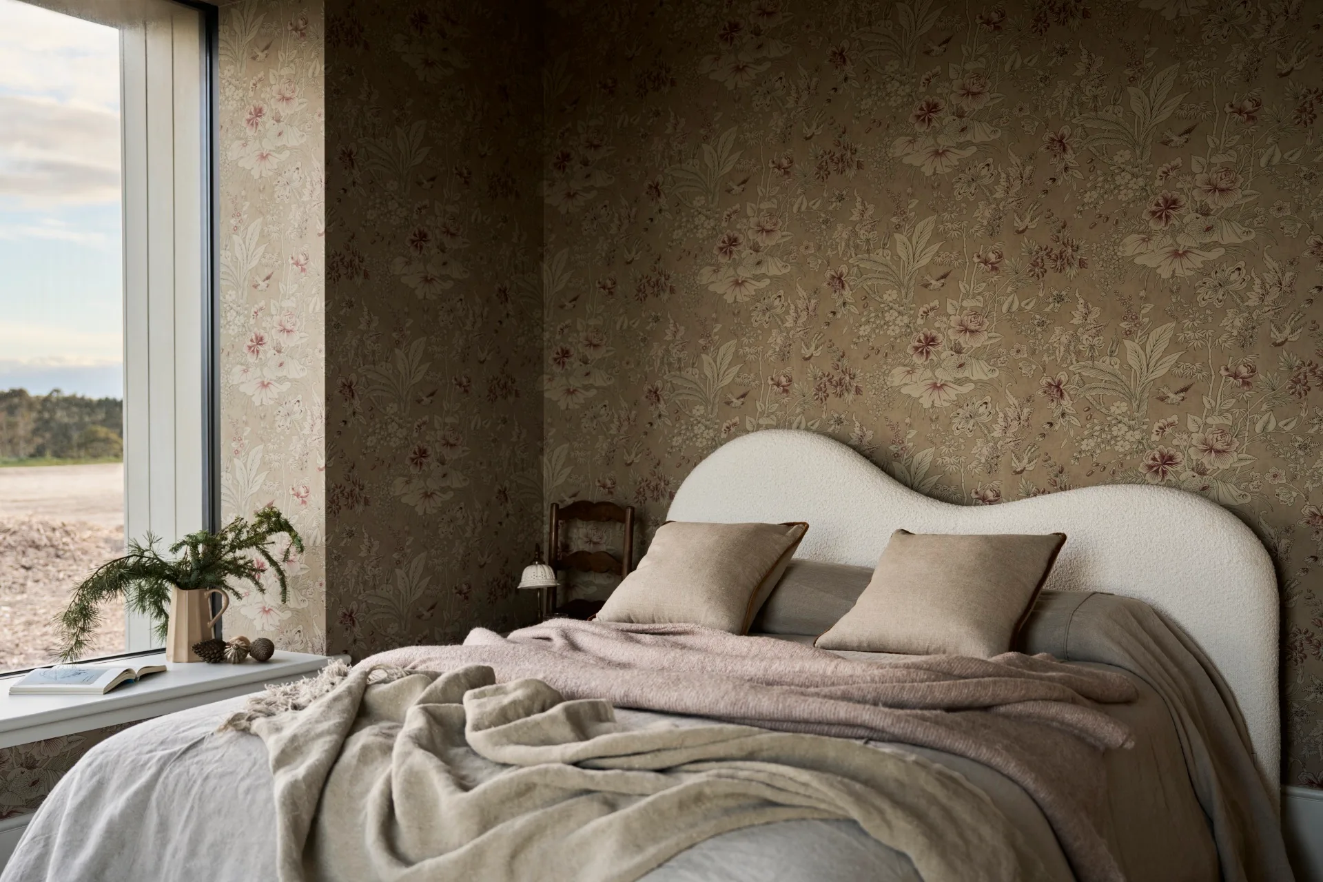 Bedroom with a brown floral wallpaper, with white curved and asymmetrical headboard, neutral bedding, books, and plants by a large window.