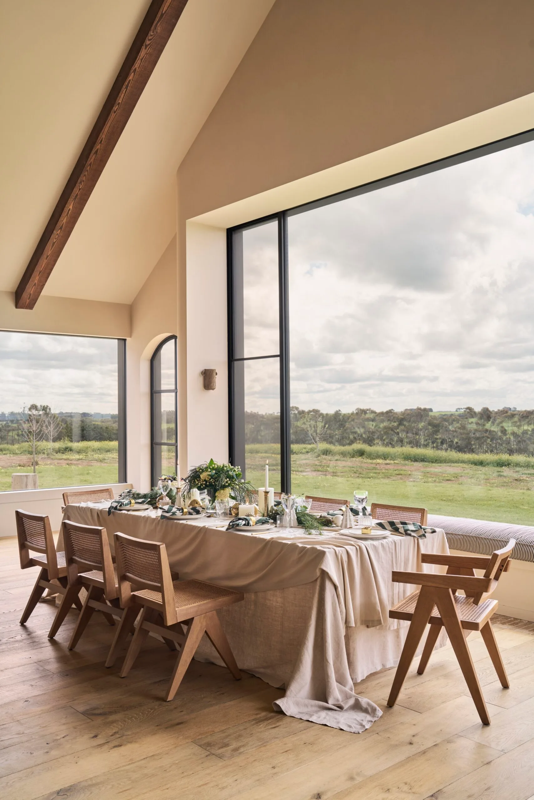 Dining table set in a modern farmhouse style open plan dining area, with large windows looking out to sweeping countryside views. Vaulted ceiling has exposed timber beams. Christmas decorations are scattered along the length of the dining table, around place settings.