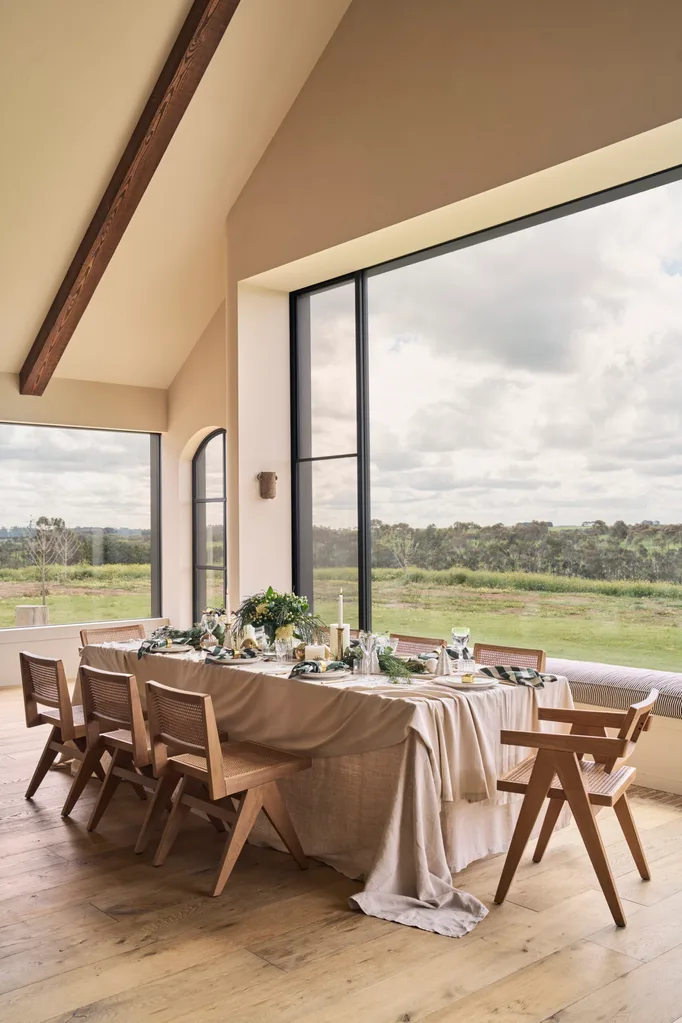 Dining table set in a modern farmhouse style open plan dining area, with large windows looking out to sweeping countryside views. Vaulted ceiling has exposed timber beams. Christmas decorations are scattered along the length of the dining table, around place settings.