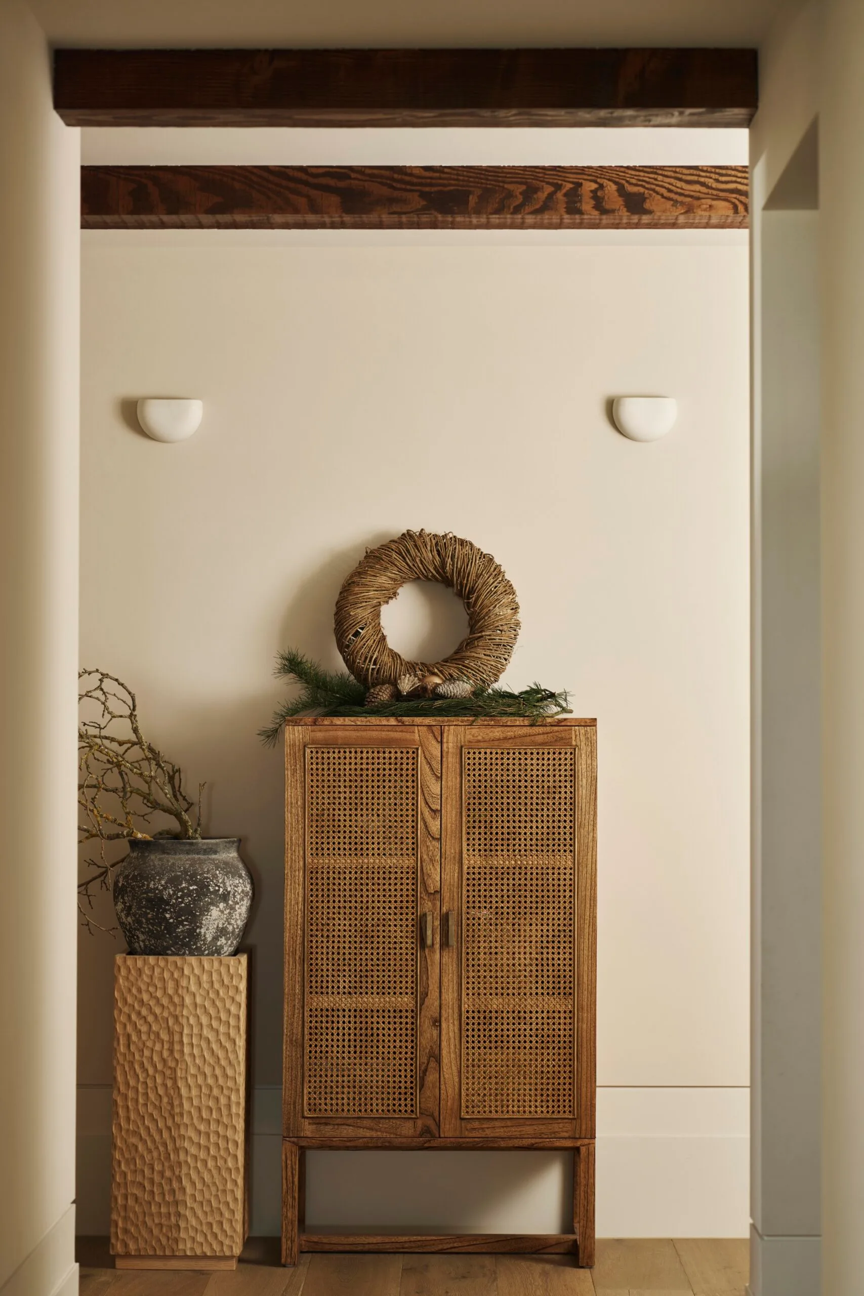 Hallway with wooden and rattan cabinet, wicker wreath, ceramic vase with branches on display, and wall lights. Exposed timber beams are near the ceiling. The style is modern farmhouse.