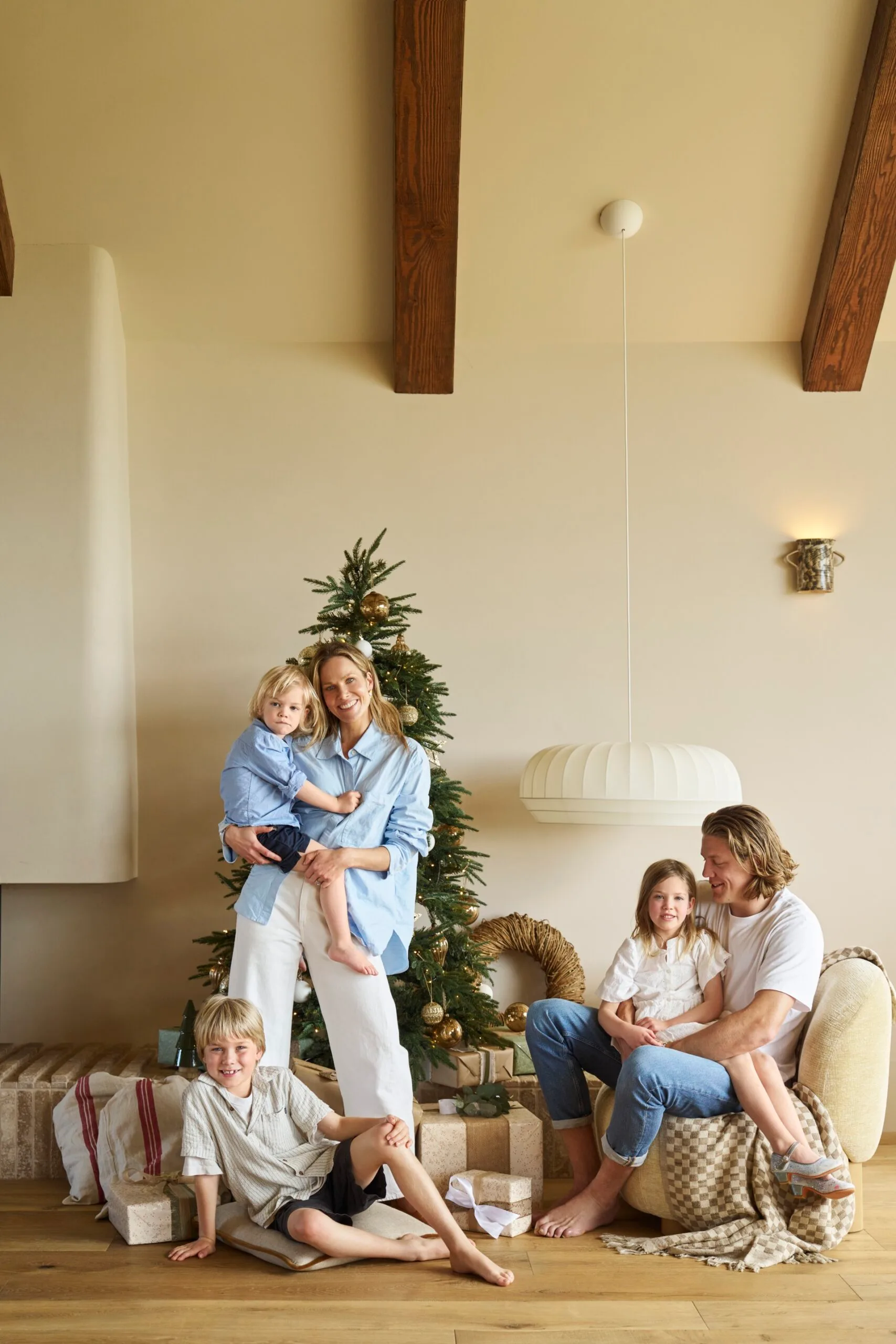 Family posing in living room, Christmas tree, two adults with three children, wrapped gifts, warm atmosphere. The style is a modern farmhouse, with vaulted ceiling and exposed timber beams. The family are Kirsten and Rhys Stanley, with their three kids.