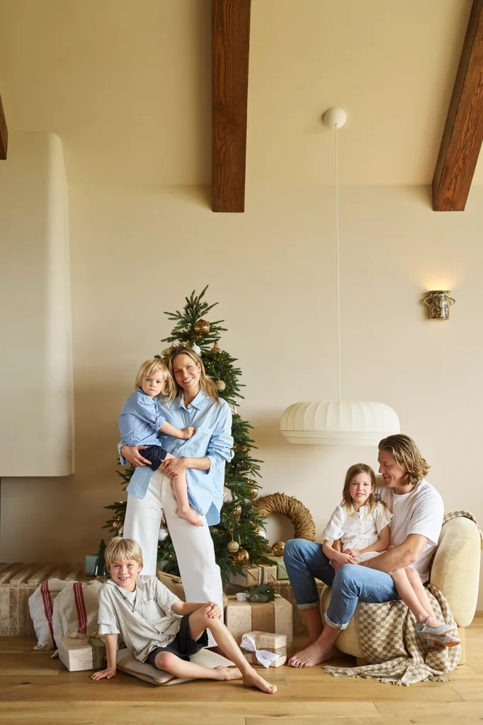 Family posing in living room, Christmas tree, two adults with three children, wrapped gifts, warm atmosphere. The style is a modern farmhouse, with vaulted ceiling and exposed timber beams. The family are Kirsten and Rhys Stanley, with their three kids.