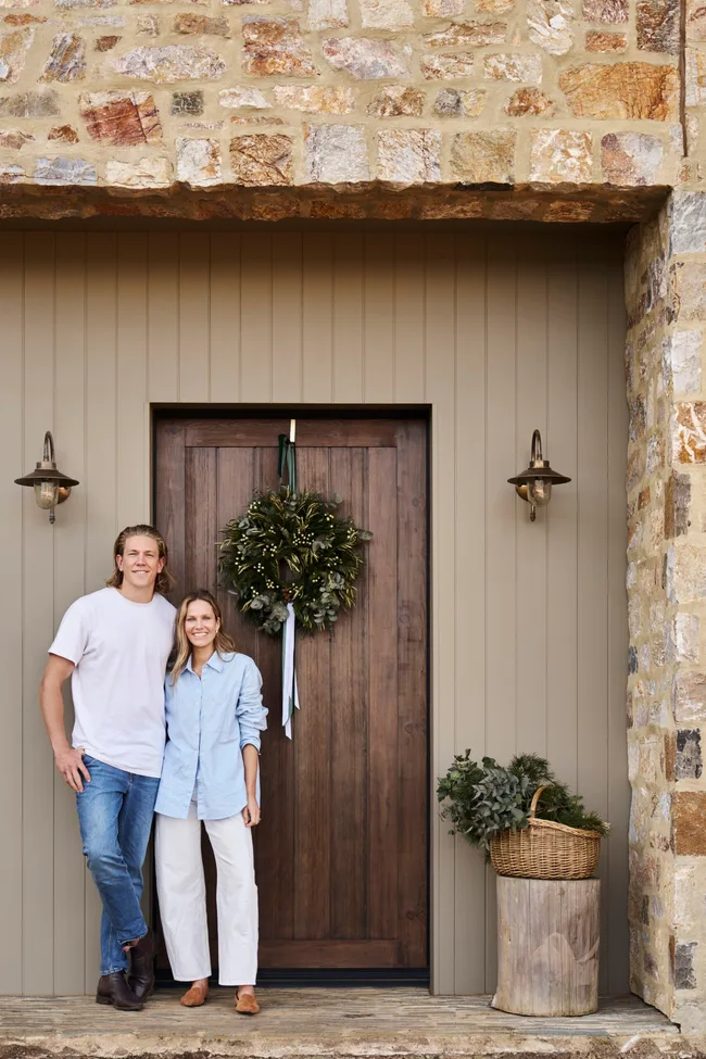 Kirsten and Rhys Stanley in front of their front door. The home is a modern farmhouse style with stone cladding, VJ panelling and a wooden door. A wreath for Christmas decorates the door.