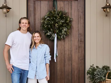 Kirsten and Rhys Stanley in front of their front door. The home is a modern farmhouse style with stone cladding, VJ panelling and a wooden door. A wreath for Christmas decorates the door.