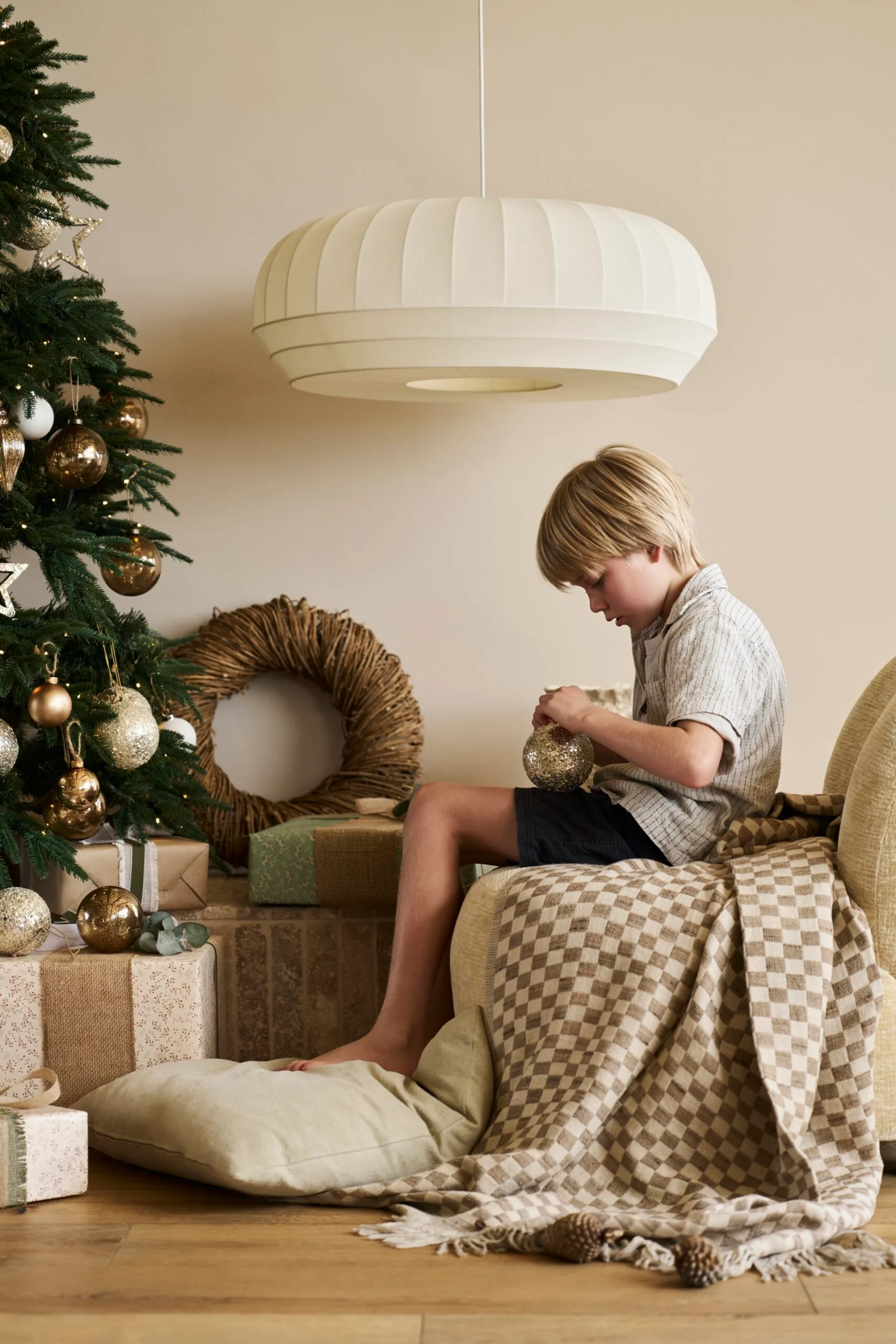 A young boy sits by a decorated Christmas tree, examining a golden ornament. A woven wreath is behind him as is a large, white pendant. The boy is the son of Kirsten and Rhys Stanley.