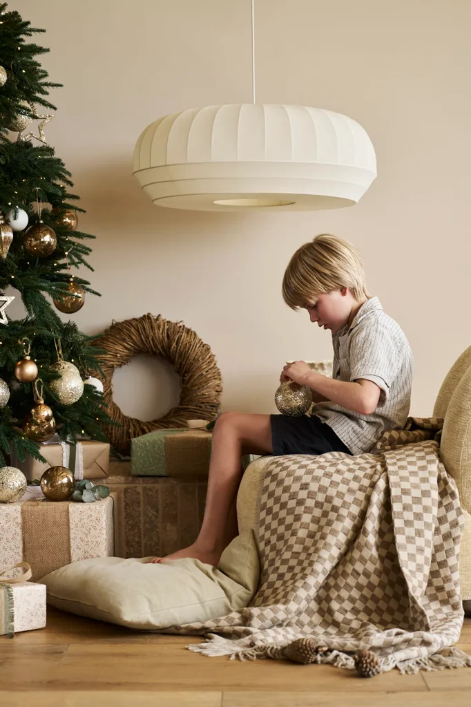 A young boy sits by a decorated Christmas tree, examining a golden ornament. A woven wreath is behind him as is a large, white pendant. The boy is the son of Kirsten and Rhys Stanley.