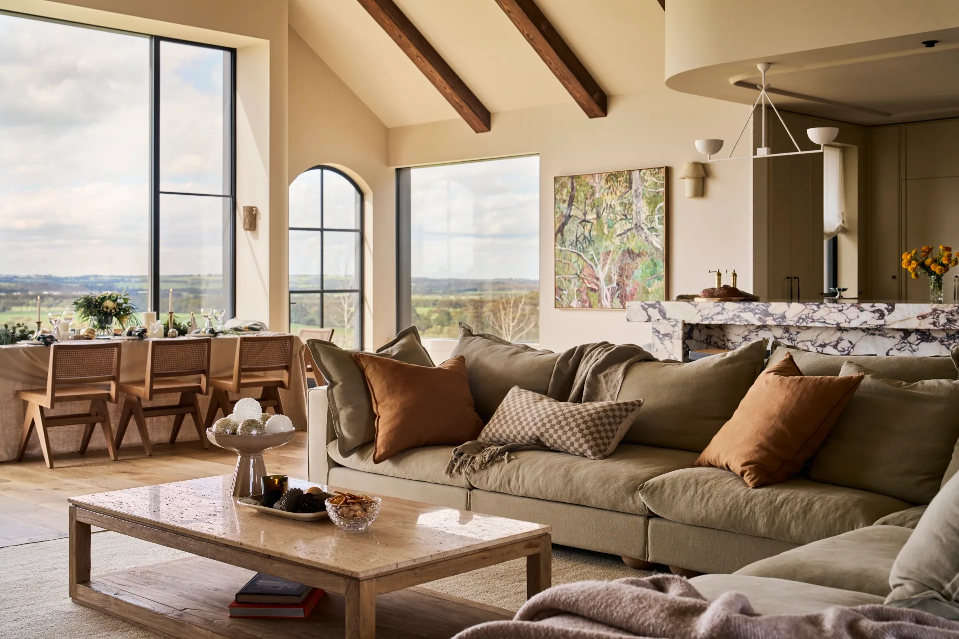 Luxurious open-plan kitchen, dining and living area. The living area is in the foreground. It has olive toned sofas, the kitchen has a marble island, and in the background large windows overlook a scenic landscape. The style is a modern farmhouse with European touches.