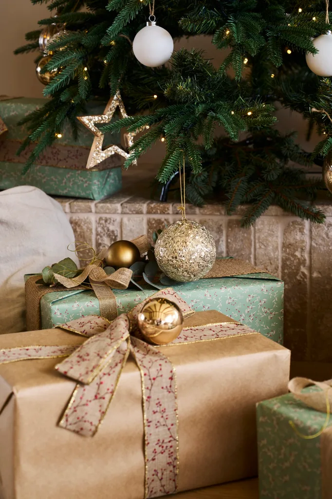 A close up photo of the bottom of a Christmas tree, decorated with warm white lighting and gold and white ornaments. Beneath the tree are wrapped Christmas presents. The wrapping paper is a mix of greens and golds, with bows in golden colours with small red floral patterns.