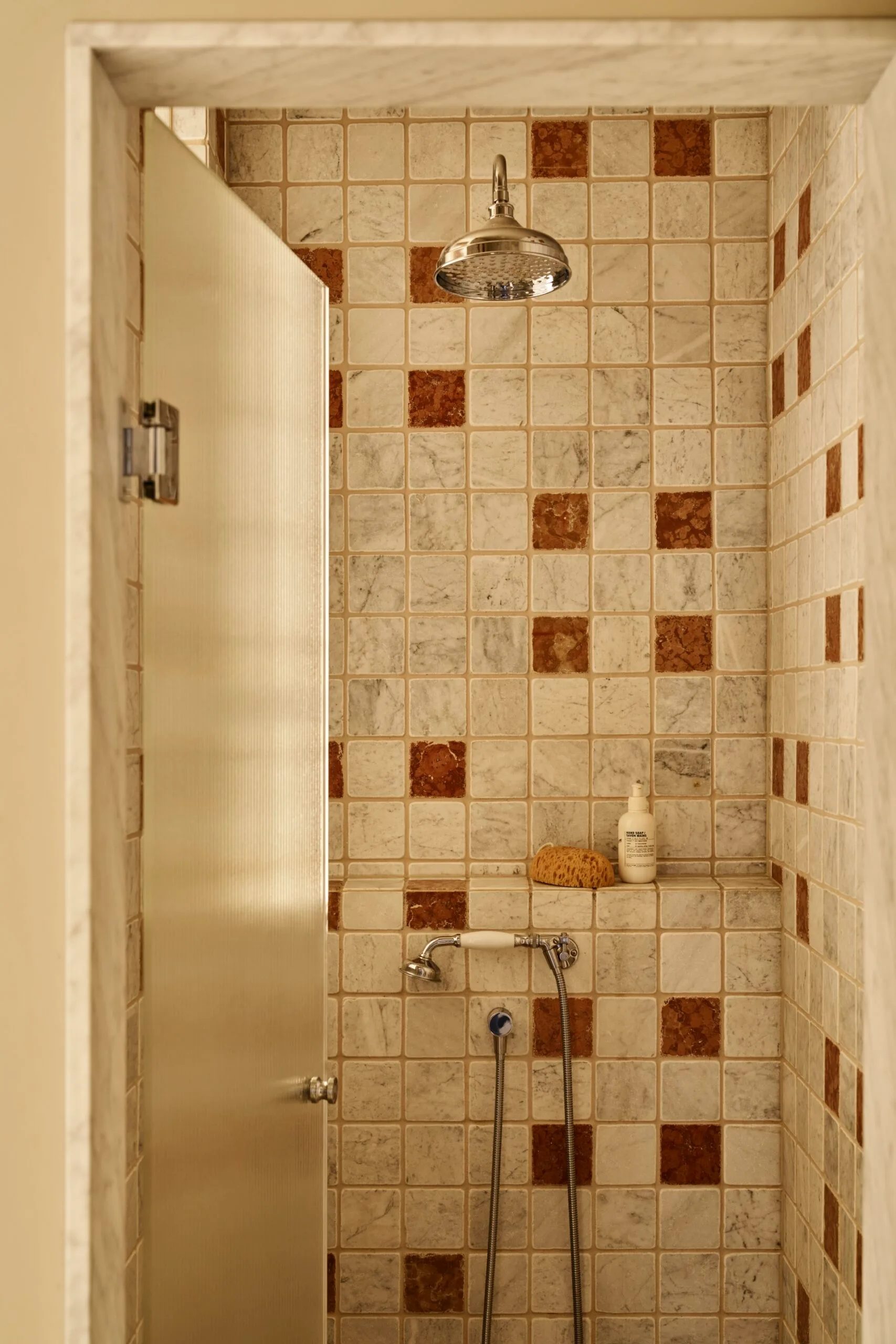 Walk-in shower with white and brown tiles, featuring a nickel showerhead and tapware, a built-in ledge with a sponge and product atop.