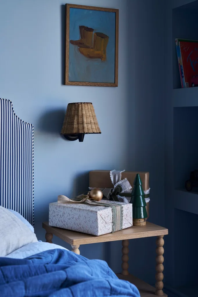The corner of a blue bedroom with striped blue and white headboard, wrapped Chritmas gifts on a table, wicker lampshade on a wall sconce, and a painting of cowboy boots on the wall. This is the bedroom of the son of Kirsten and Rhys Stanley.