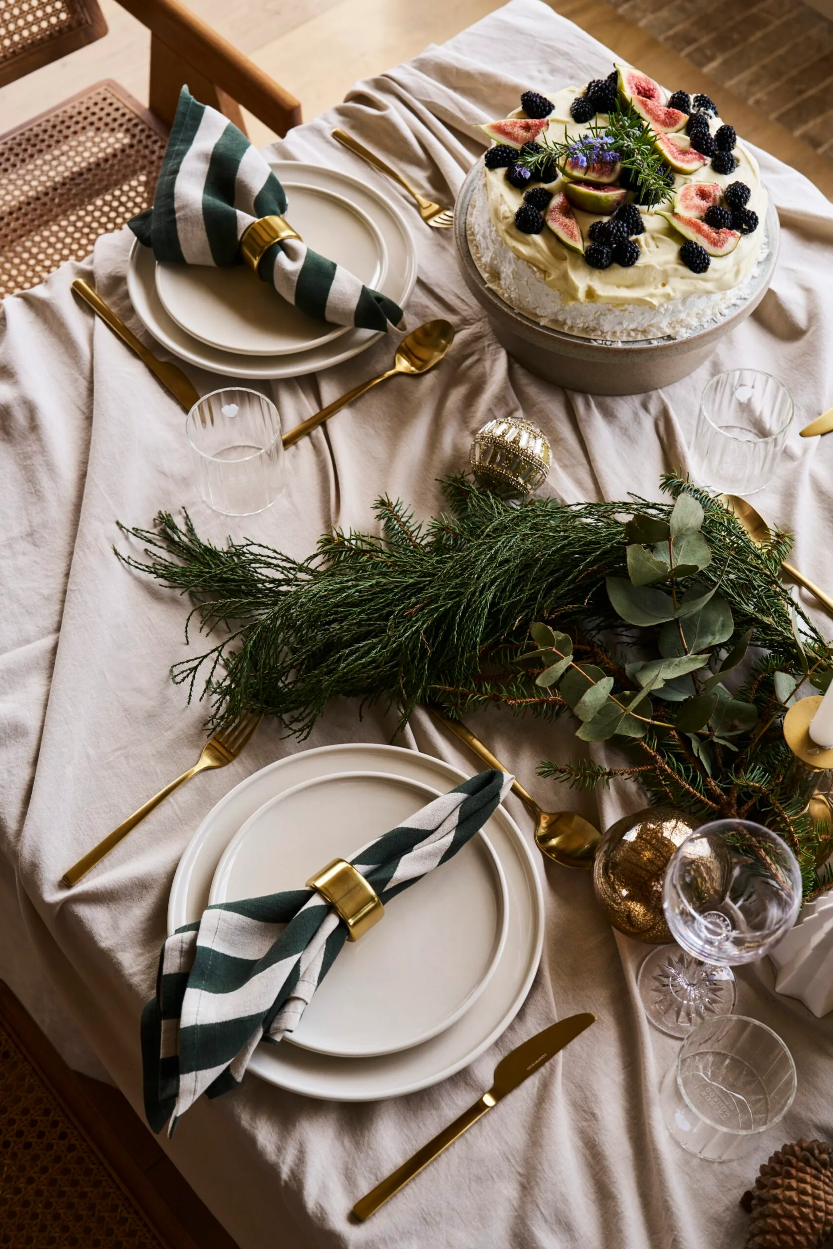 Elegant holiday table setting with neutral toned dinnerware. candles and brass candlesticks, greenery from pine trees, and decorative white trees with gold stars on top. These are all atop neutral table linen. A timber dining chair, with rattan seat and back, is visible. A pavlova is also on the table, while green and white napkins are placed on top of plates with a brass napkin ring.