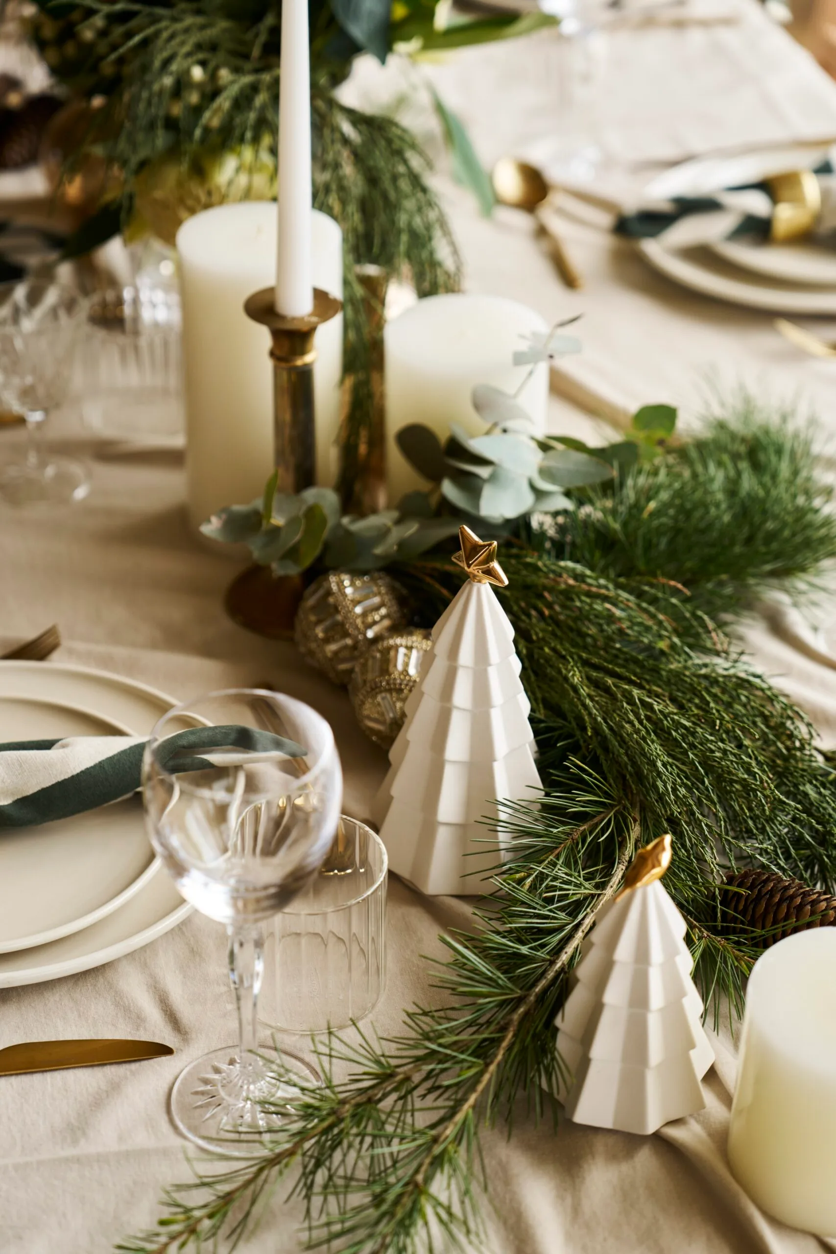 Elegant holiday table setting with neutral toned dinnerware. candles and brass candlesticks, greenery from pine trees, and decorative white trees with gold stars on top. These are all atop neutral table linen.