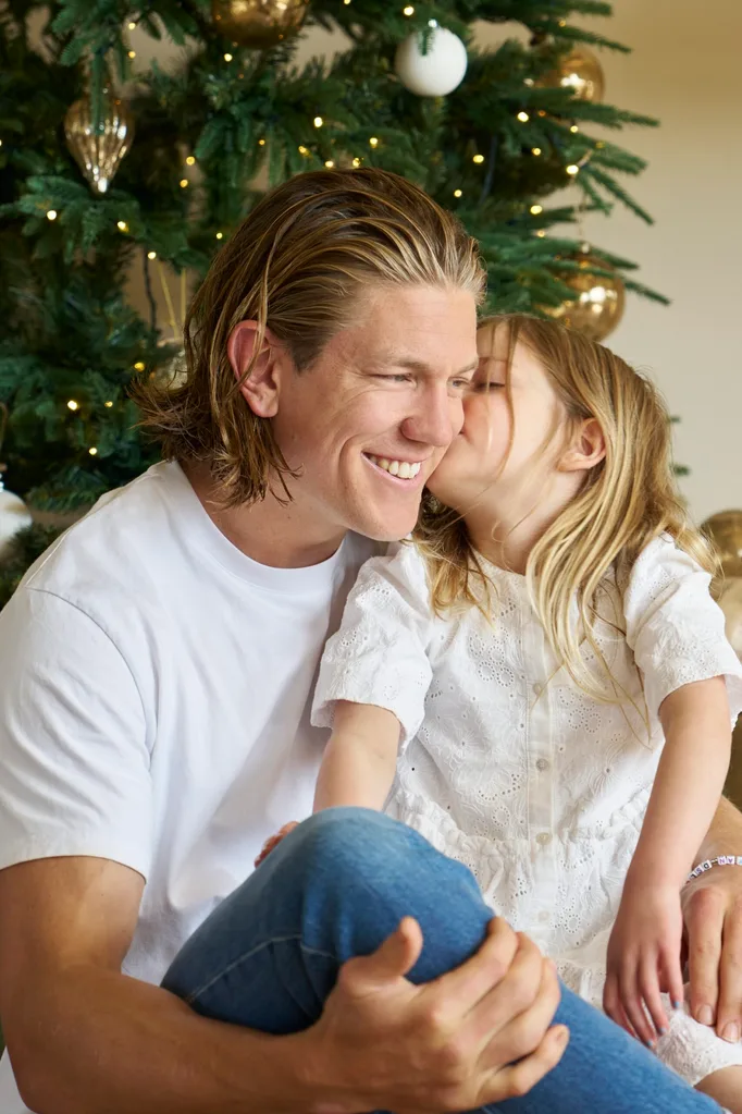 A girl in a white dress kisses the cheek of her father. Both have blond hair and the dad is dressed in a white t-shirt and blue jeans. They sit in front of a decorated Christmas tree. The father is AFL star Rhys Stanley.