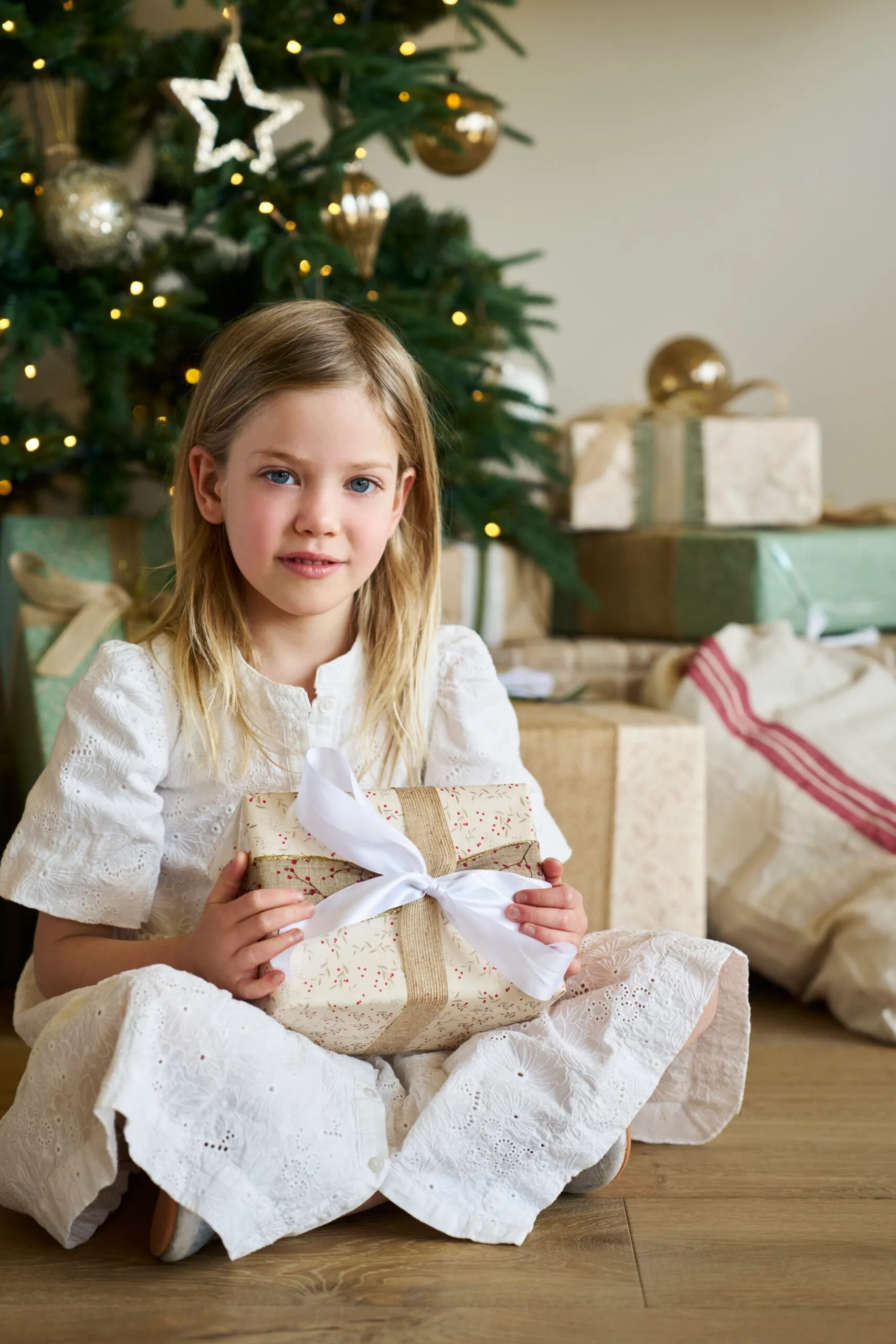 Girl in white dress holding a gift in front of a decorated Christmas tree with wrapped presents underneath. The wrapping paper is cream with small red floral printed on it. The girl is the daughter of Kirsten and Rhys Stanley.