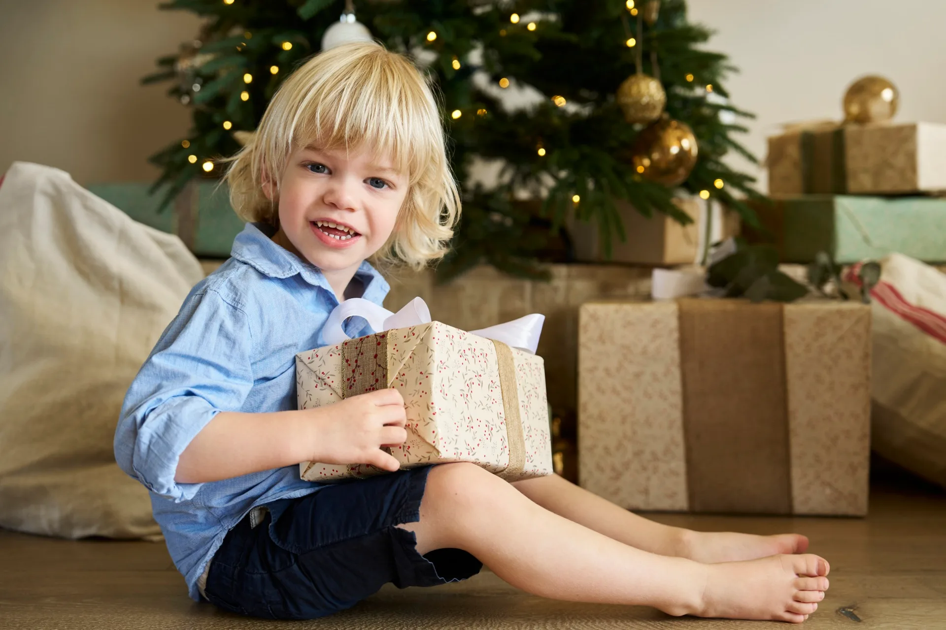 Young boy with blond hair holding a gift in front of a Christmas tree with presents underneath. The wrapping paper is cream with small red floral printed on it. The boy is the son of Kirsten and Rhys Stanley.