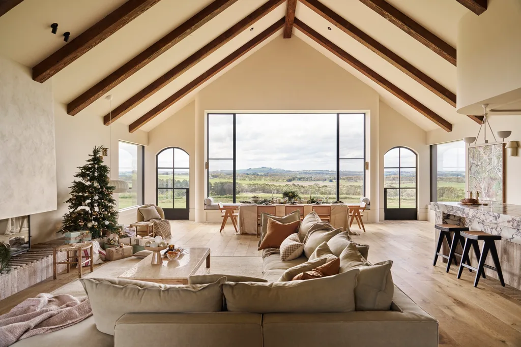 Spacious living room with high vaulted ceilings and exposed timber beams, large windows, cozy furniture, Christmas tree, and a scenic countryside view. The style of the home is modern farmhouse with a European twist.