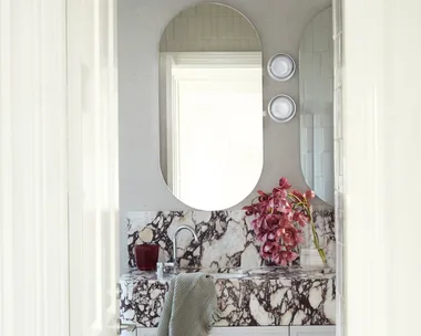 A bathroom with a marble sink, oval mirrors, pink flowers, and a gray towel hanging on the counter.