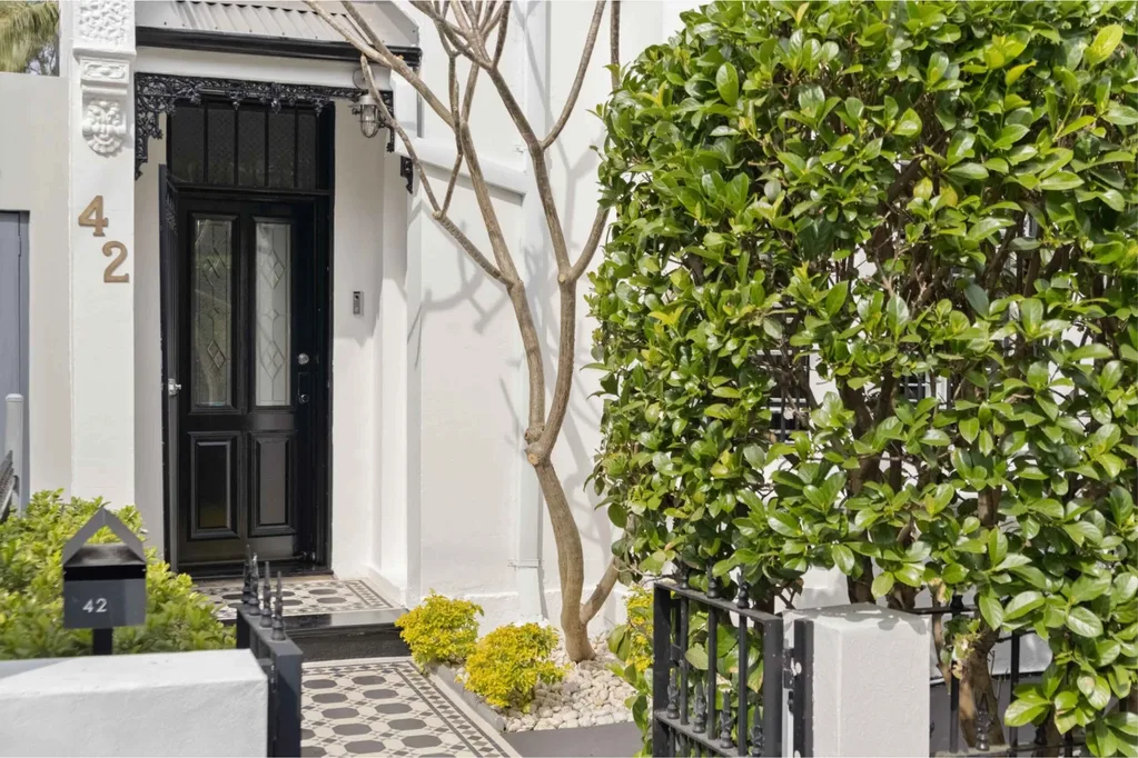 Front door and porch of a white and black Victorian house terrace-style house with a garden hedge and tesselated tiles.