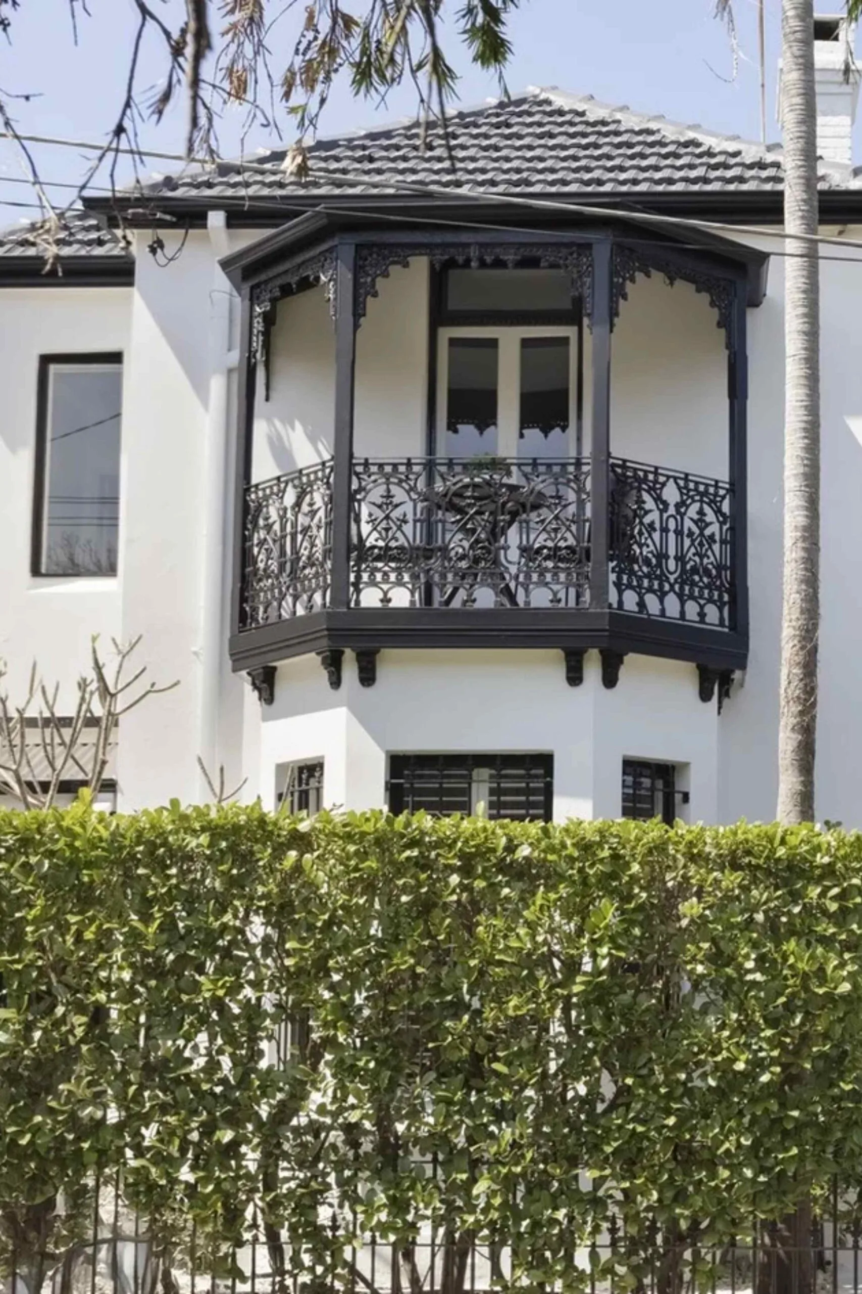 Front of a white Victorian terrace-style house with black fretwork and a garden hedge.