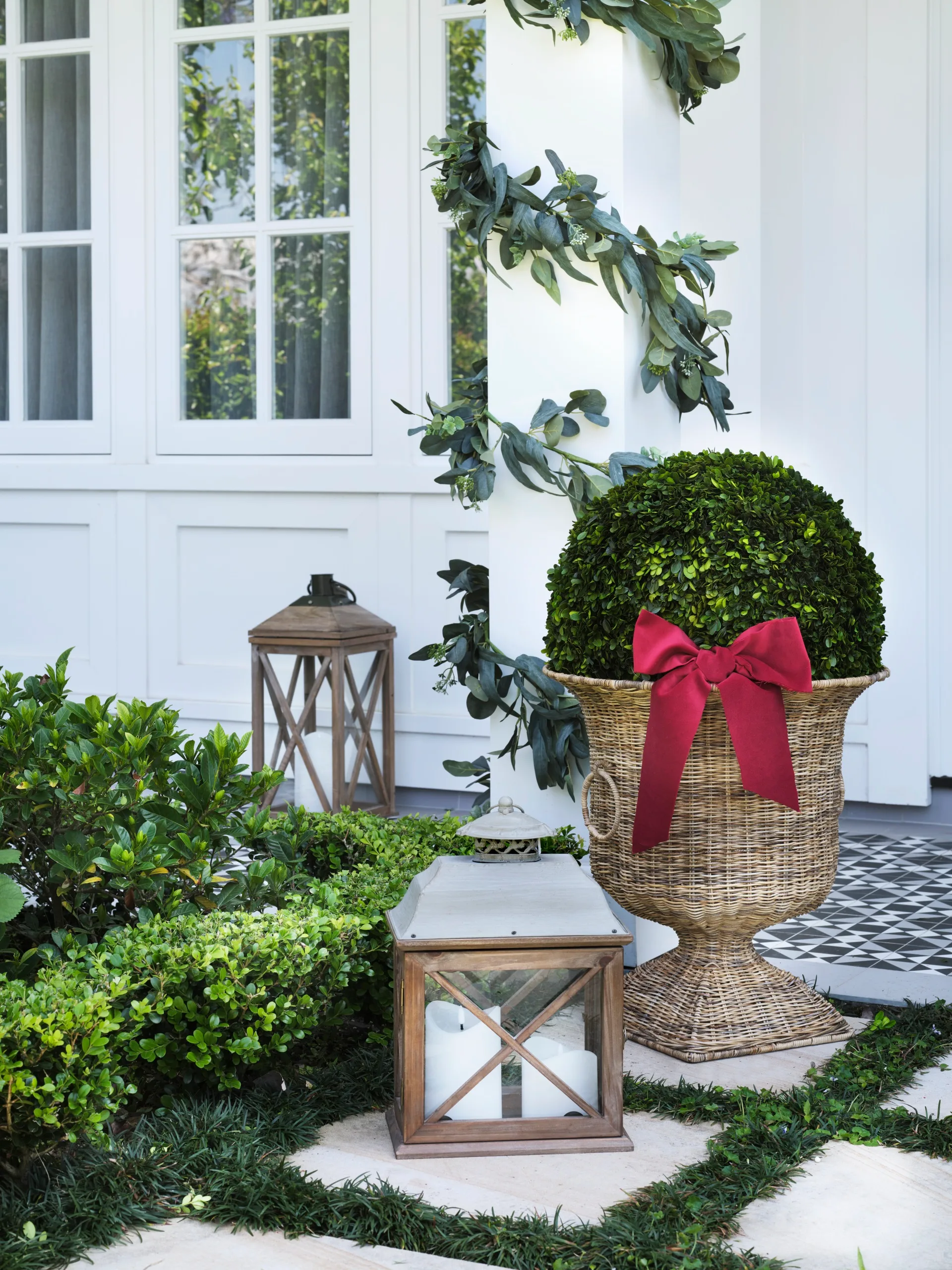 white home facade with Christmas decorations of potted topiary in wicker urn and hurricane lanterns in front of columns decorated with christmas garlands