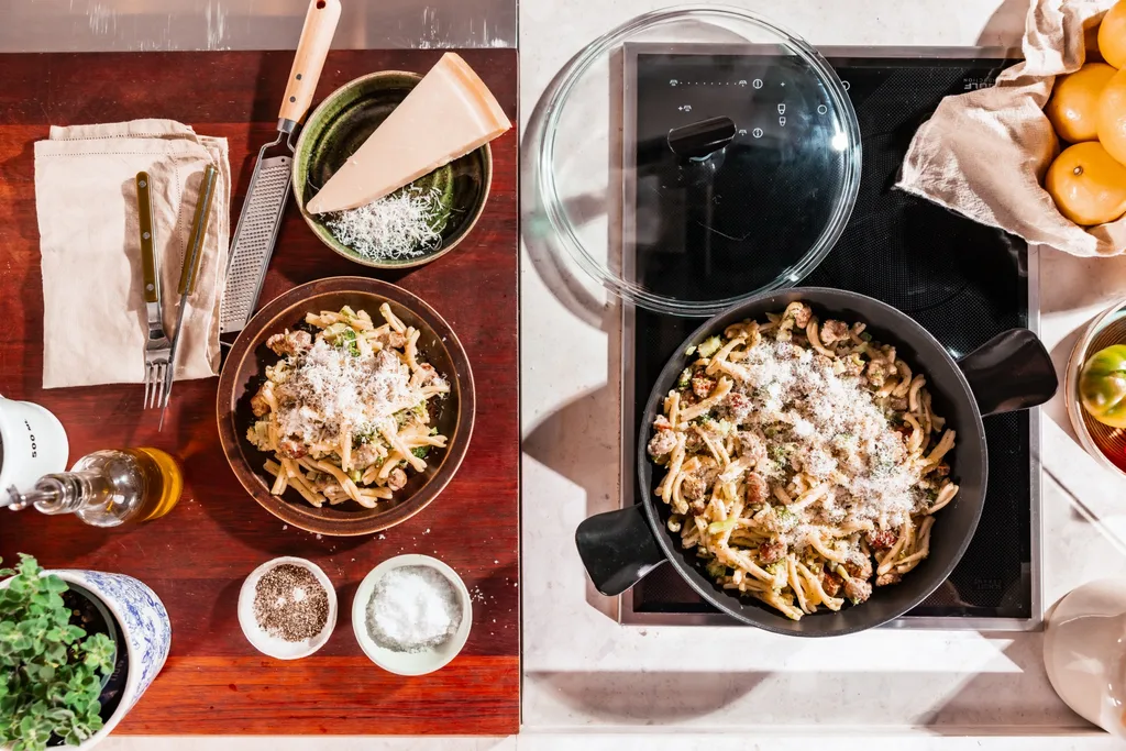 A kitchen benchtop as seen from above. A sauteuse has pasta that has finished cooking in it while a chopping board beside this has a bowl filled with the pasta, a bowl with a triangle of cheese that has been partially grated plus a pot with a plant and a bottle of olive oil.