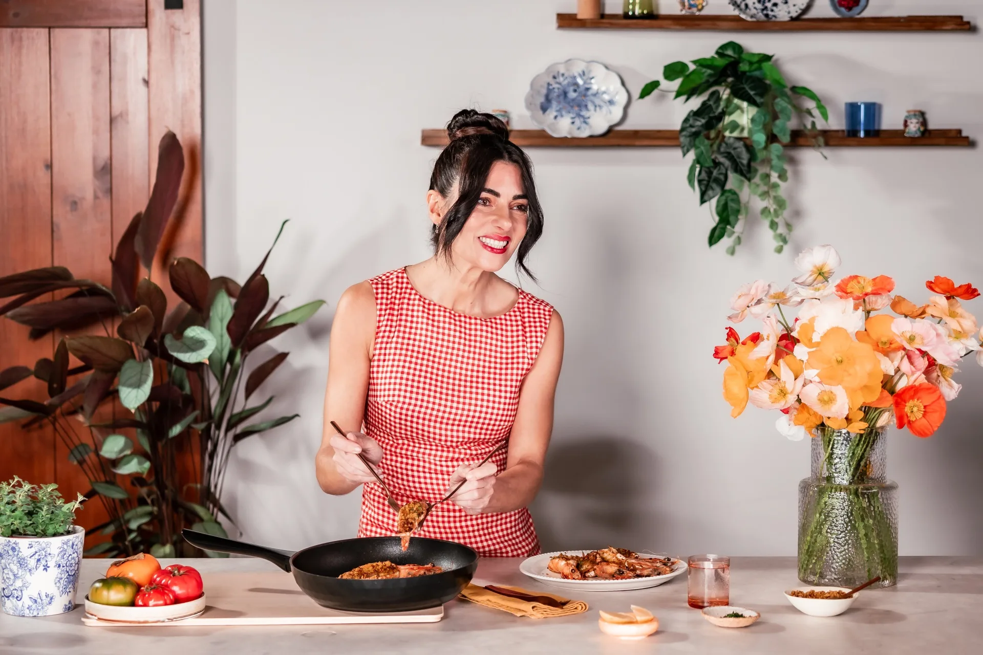Actress and cook Silvia Colloca in a kitchen with a red and white gingham dress on. She is spooning a prawn dish out of a saucepan and on to a plate. A few potted plants and flowers in vases are positioned around her.