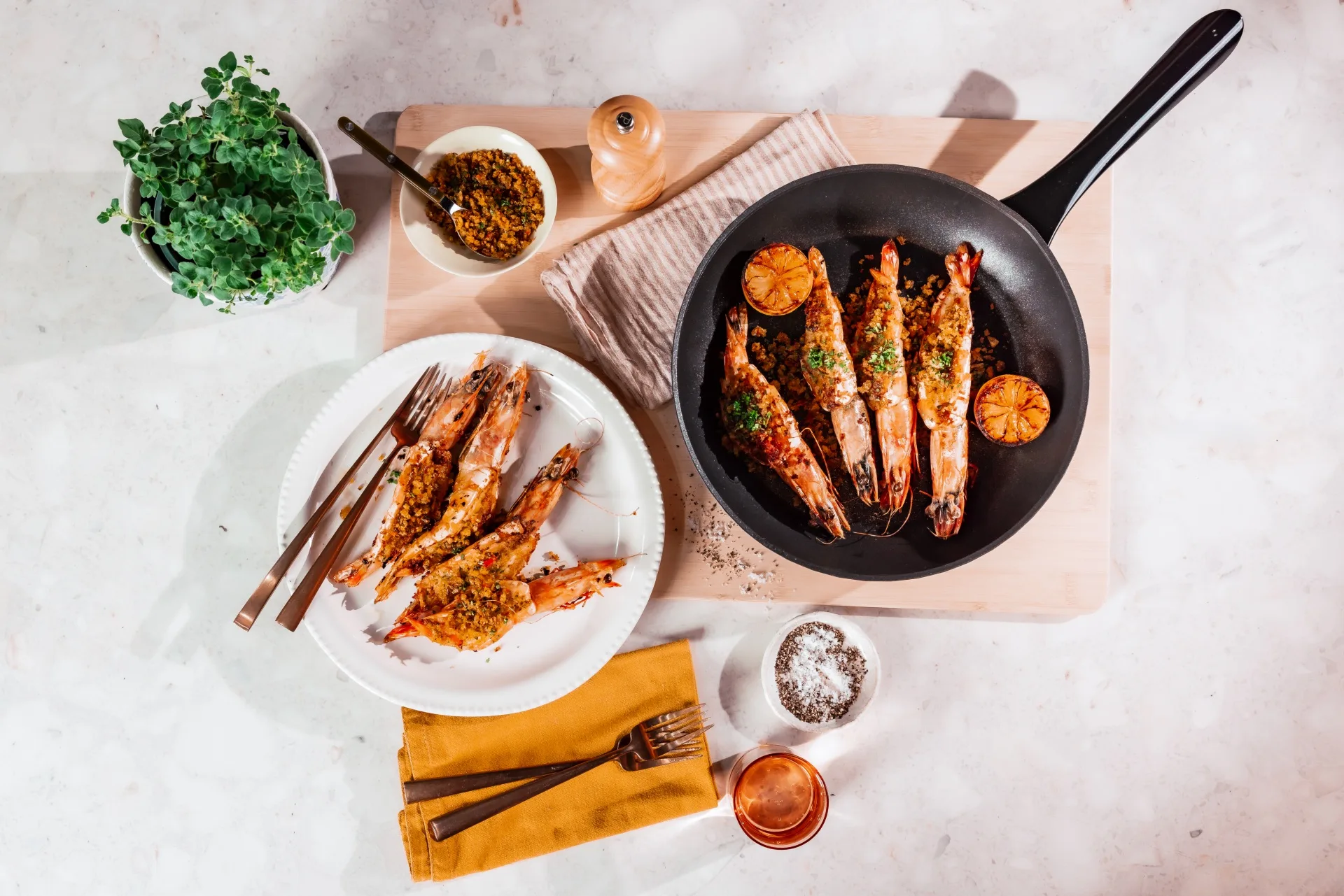 A flatlay image of a kitchen benchtop. A saucepan is atop a chopping board, filled with large prawns that are also placed on a dinner plate beside the pan. Cutlery is on a mustard toned napkin.
