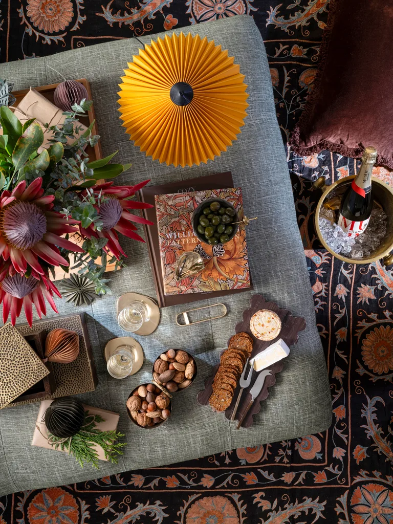 Festive ottoman styled and decorated with a floral rug, cheese, nuts, a lamp, champagne and gifts.