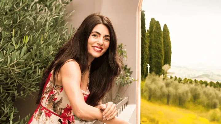 Actress, opera singer and cook Silvia Colloca has long dark hair, red lips and a red dress. She leans over a balcony railing with an olive tree behind her and vast landscape visible beyond.