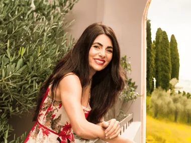 Actress, opera singer and cook Silvia Colloca has long dark hair, red lips and a red dress. She leans over a balcony railing with an olive tree behind her and vast landscape visible beyond.