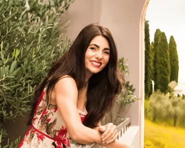 Actress, opera singer and cook Silvia Colloca has long dark hair, red lips and a red dress. She leans over a balcony railing with an olive tree behind her and vast landscape visible beyond.