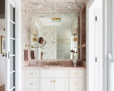 A vanity in an ensuite, situated in a spacious bedroom suite. Pink toned marble graces the vanity benchtop and a niche on the wall, which has open shelves also made from this pink marble. The joinery is white, complemented by a white wallpaper with a dusty rose botanic pattern. Floor tiles are white marble.