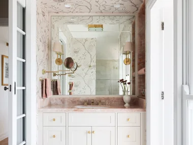 A vanity in an ensuite, situated in a spacious bedroom suite. Pink toned marble graces the vanity benchtop and a niche on the wall, which has open shelves also made from this pink marble. The joinery is white, complemented by a white wallpaper with a dusty rose botanic pattern. Floor tiles are white marble.