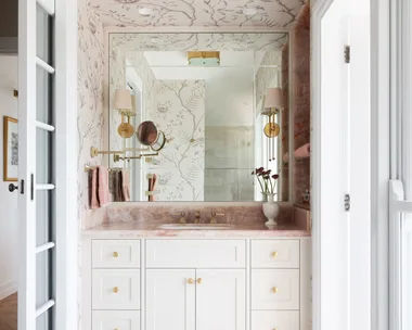 A vanity in an ensuite, situated in a spacious bedroom suite. Pink toned marble graces the vanity benchtop and a niche on the wall, which has open shelves also made from this pink marble. The joinery is white, complemented by a white wallpaper with a dusty rose botanic pattern. Floor tiles are white marble.