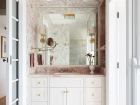 A vanity in an ensuite, situated in a spacious bedroom suite. Pink toned marble graces the vanity benchtop and a niche on the wall, which has open shelves also made from this pink marble. The joinery is white, complemented by a white wallpaper with a dusty rose botanic pattern. Floor tiles are white marble.