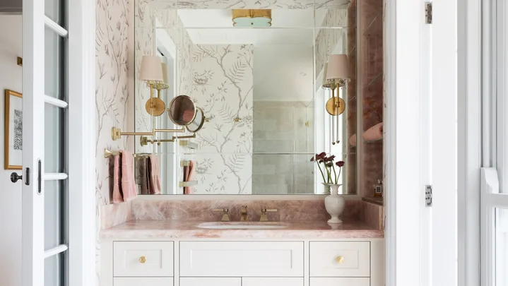 A vanity in an ensuite, situated in a spacious bedroom suite. Pink toned marble graces the vanity benchtop and a niche on the wall, which has open shelves also made from this pink marble. The joinery is white, complemented by a white wallpaper with a dusty rose botanic pattern. Floor tiles are white marble.