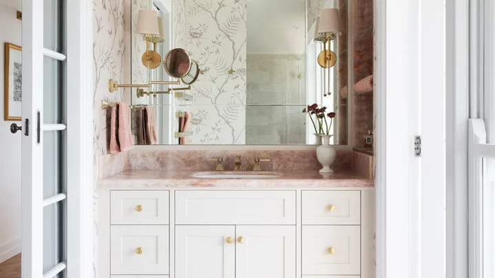 A vanity in an ensuite, situated in a spacious bedroom suite. Pink toned marble graces the vanity benchtop and a niche on the wall, which has open shelves also made from this pink marble. The joinery is white, complemented by a white wallpaper with a dusty rose botanic pattern. Floor tiles are white marble.