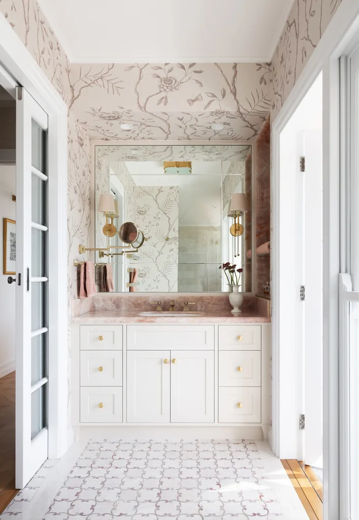 A vanity in an ensuite, situated in a spacious bedroom suite. Pink toned marble graces the vanity benchtop and a niche on the wall, which has open shelves also made from this pink marble. The joinery is white, complemented by a white wallpaper with a dusty rose botanic pattern. Floor tiles are white marble.
