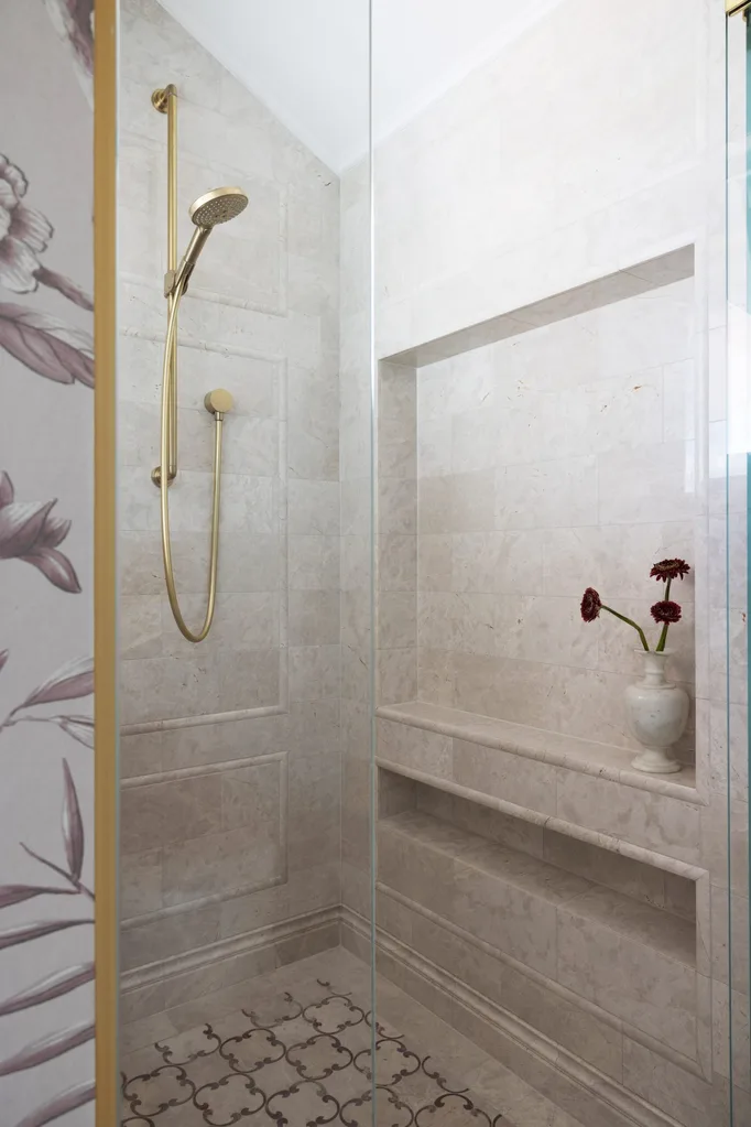 A large shower inside an ensuite, which is part of a bedroom suite. Blush toned marble tiles grace the walls and shower niche while brass hardware comprises the showerhead.