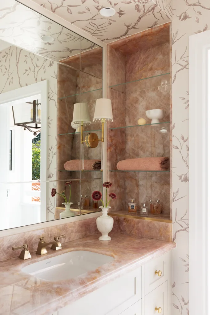 A vanity in an ensuite, situated in a spacious bedroom suite. Pink toned marble graces the vanity benchtop and a niche on the wall, which has open shelves also made from this pink marble. The joinery is white, complemented by a white wallpaper with a dusty rose botanic pattern.