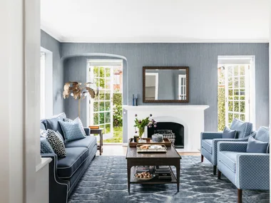 Cozy living room with blue sofa, armchairs, fireplace, coffee table, large mirror, and open French doors to garden.