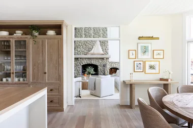 Cozy living room with stone fireplace, white sofas, wooden dining table, and framed artwork on cream walls.