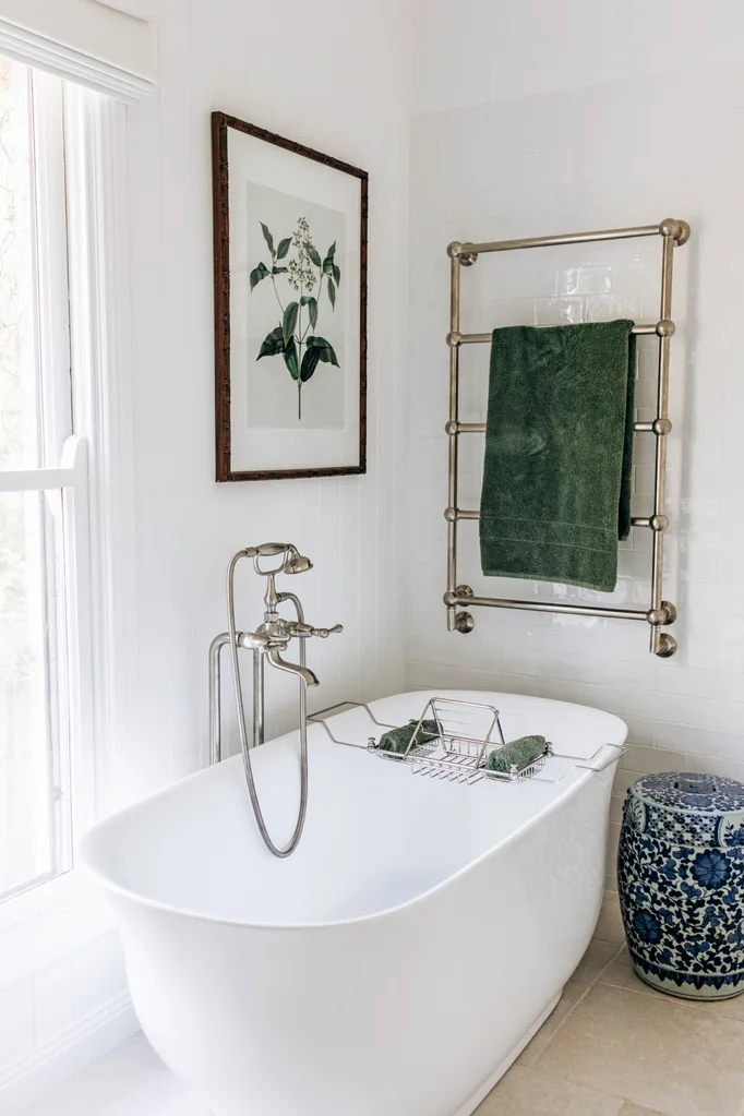 A white country cottage bathroom with green details, such as a botanical artwork and a green towel hanging on platinum towel rail. A freestanding bathtub is next to a ginger jar style stool.