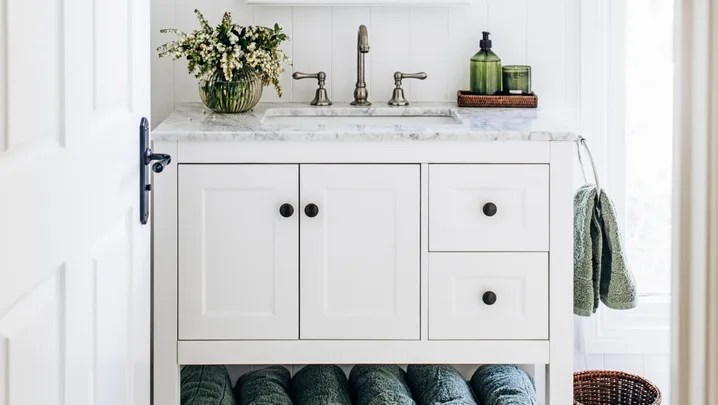A white country bathroom with green decorative details such as rolled green towels and green hand soap. Two lamps are positioned on either side of the arched mirror, above a freestanding vanity with shaker cabinets and a marble top.